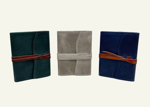 Three closed leather journals in dark green, taupe, and navy blue with contrasting leather wraparound cords.