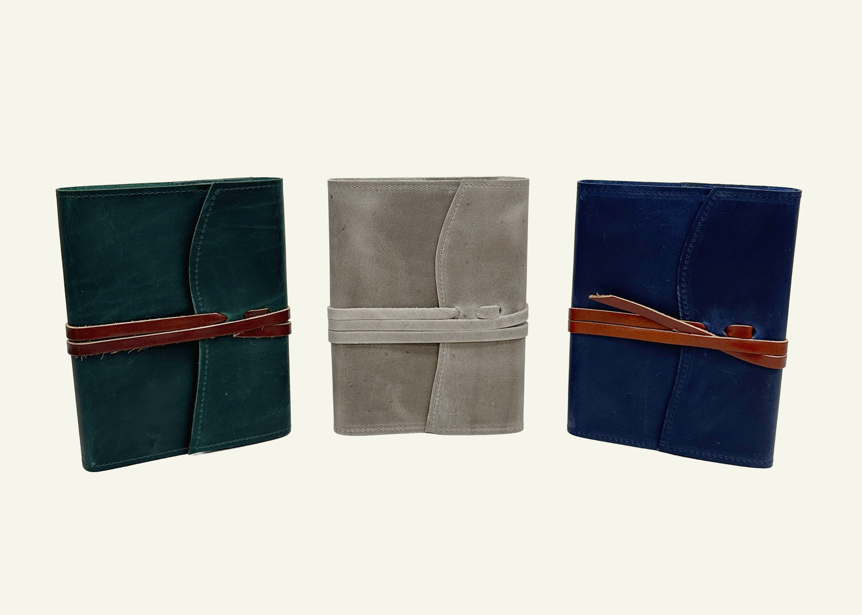 Three closed leather journals in dark green, taupe, and navy blue with contrasting leather wraparound cords.